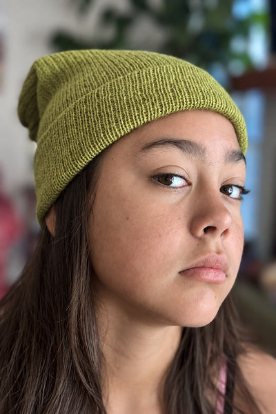 A close-up portrait of a person wearing a ribbed knit beanie hat in a muted olive green color, with a softly blurred indoor background.