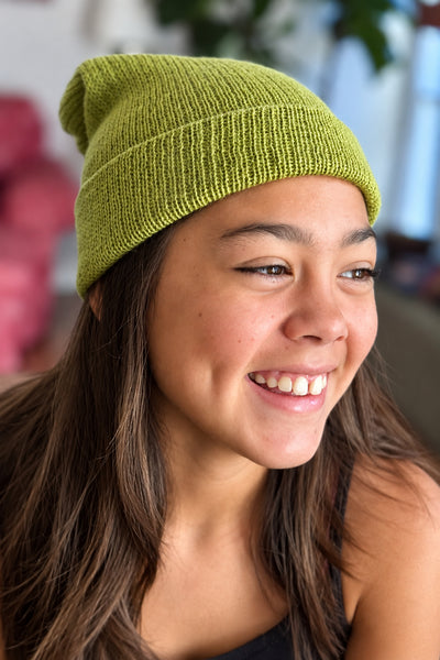 A smiling young woman wearing a textured olive green knit beanie hat, shown in a close-up portrait.