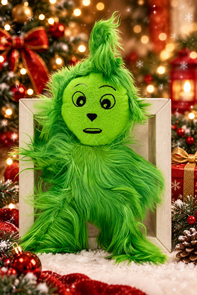A green Grinch-style plush toy with long shaggy hair and a round embroidered face, posed in front of a white frame on a light gray background.