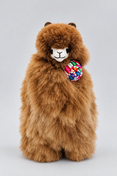 A fluffy brown alpaca plush toy holding a andean flower, photographed against a plain light gray studio background.